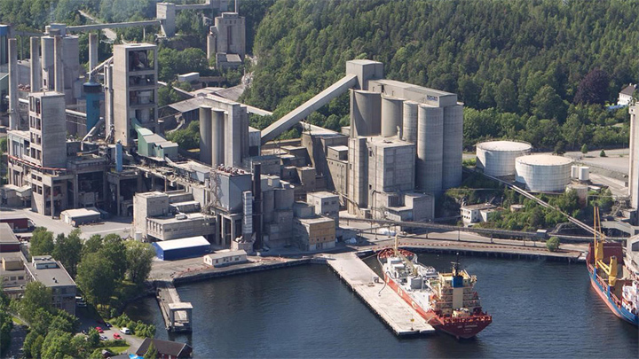 Aerial view of the Brevik CCS facility in Norway, showing a large cement plant with silos, industrial buildings, and docked cargo ships beside a fjord, surrounded by forested hills.