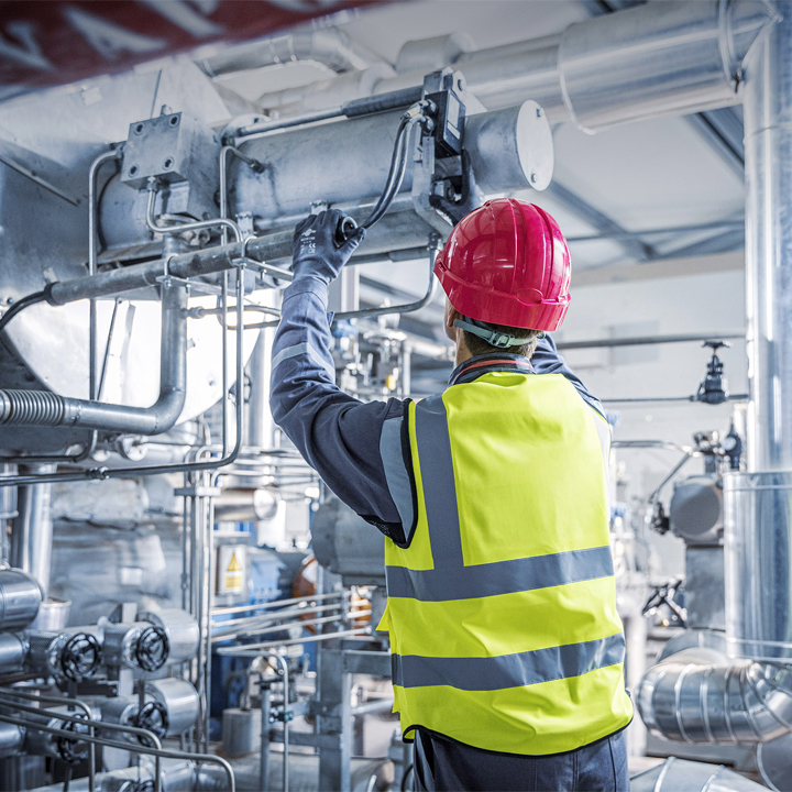 Worker in high-visibility vest and red hard hat inspecting industrial machinery with pipes and valves
