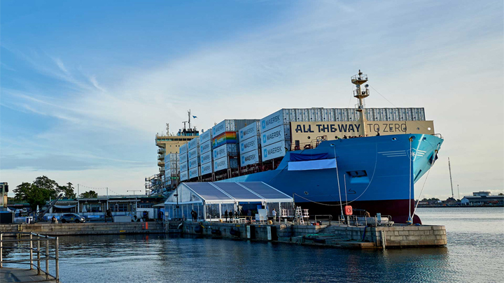 Large teal cargo ship, Laura Maersk, docked at a port beside a pier, with containers and a nearby tent structure visible.