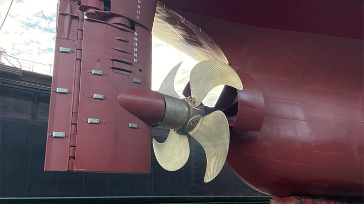 Close-up of a ship’s stern showing a red rudder, four-blade propeller, and draft markings on the hull