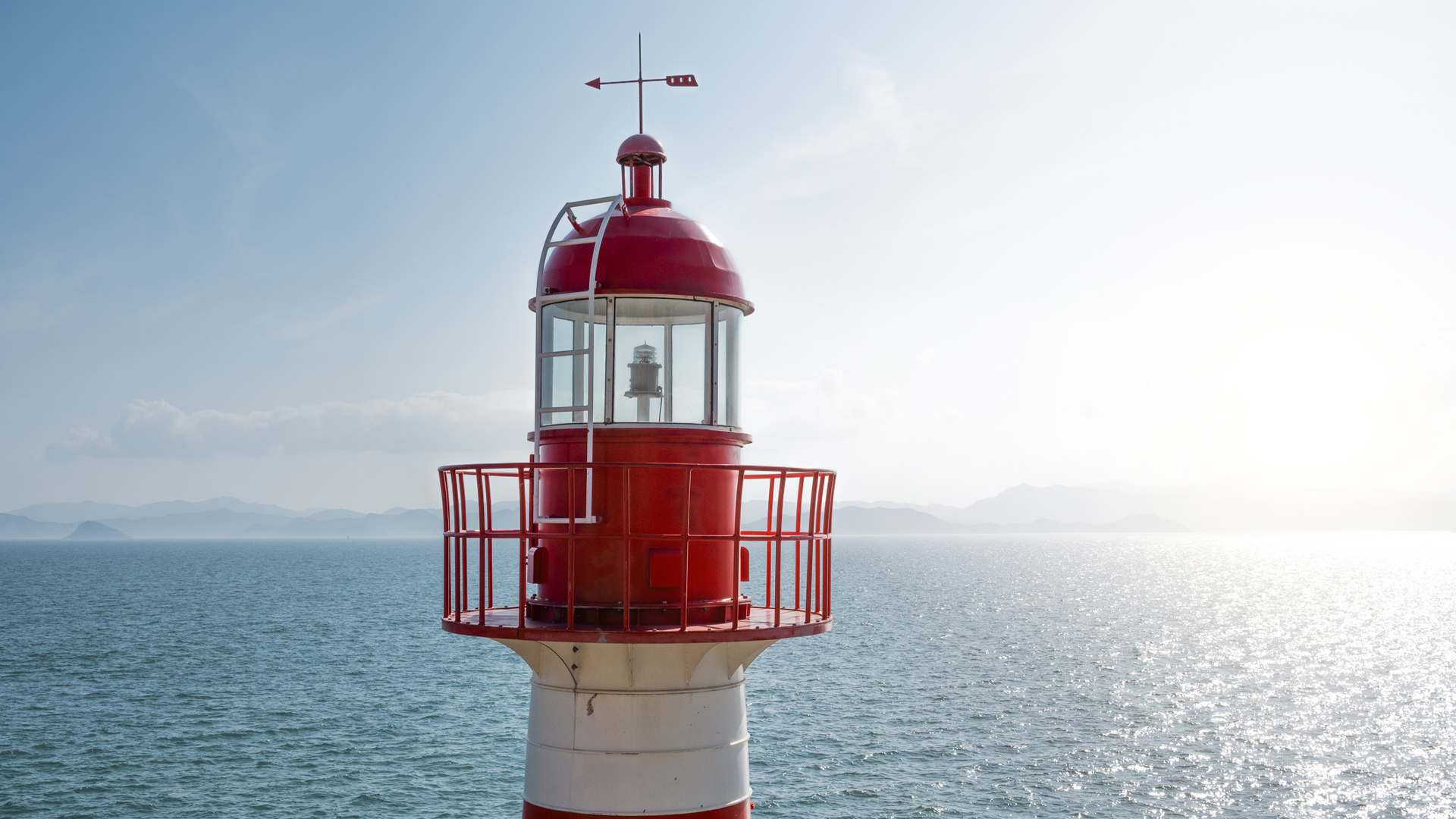Red lighthouse standing at the edge of the sea, with calm waters and distant mountains under a bright sky