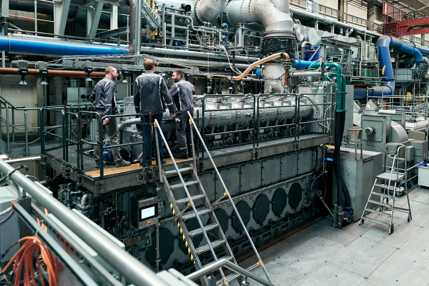 Three engineers standing on a platform inspecting a large industrial engine with pipes and valves in a factory setting